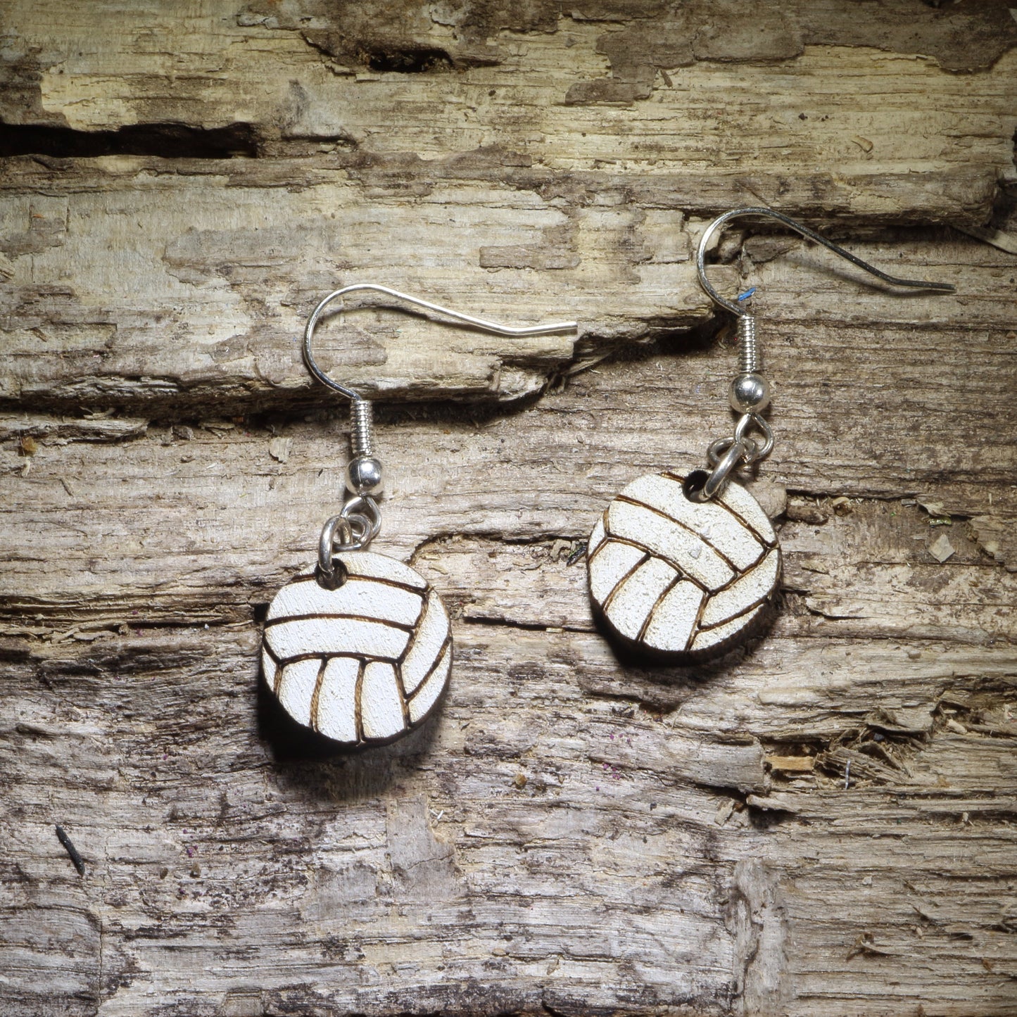 Pair of hand-painted white volleyball wooden dangle earrings featuring a classic volleyball panel design, displayed on rustic weathered wood