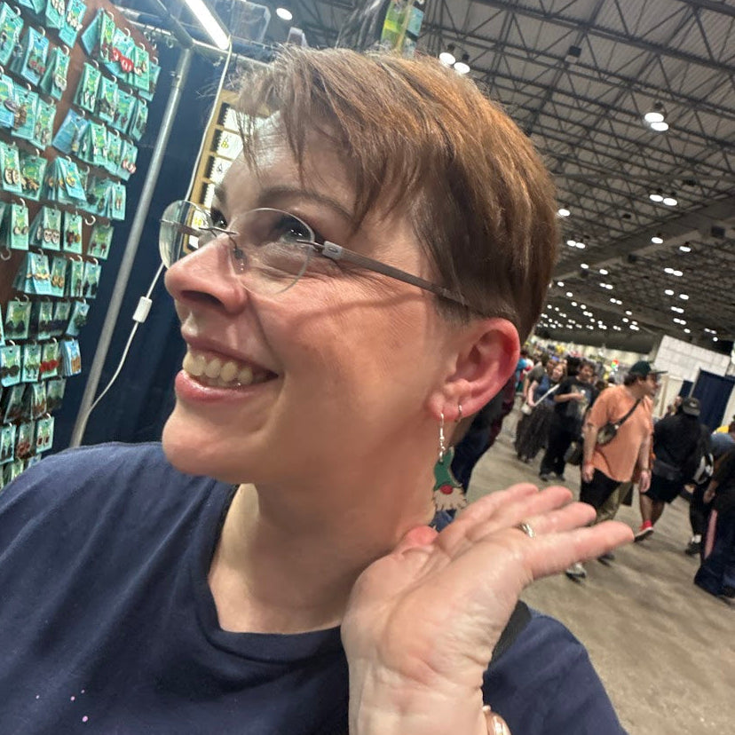 Smiling woman modeling hand-painted Garden Gnome Wooden Dangle Earrings | Whimsical Nature Jewelry Giftat a convention booth, lightweight whimsical jewelry by Cate’s Concepts