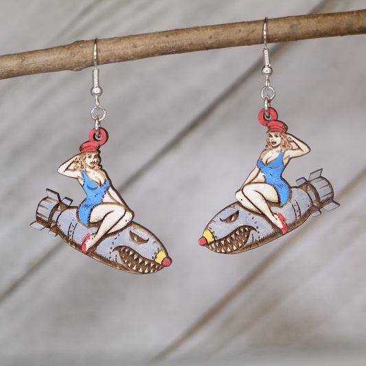 Close-up of hand-painted wooden pin-up bomber girl dangle earrings featuring vintage nose art style woman riding cartoon bomb with red hat and blue outfit and hypoallergenic sterling silver hooks, retro WWII inspired statement jewelry worn on ear
