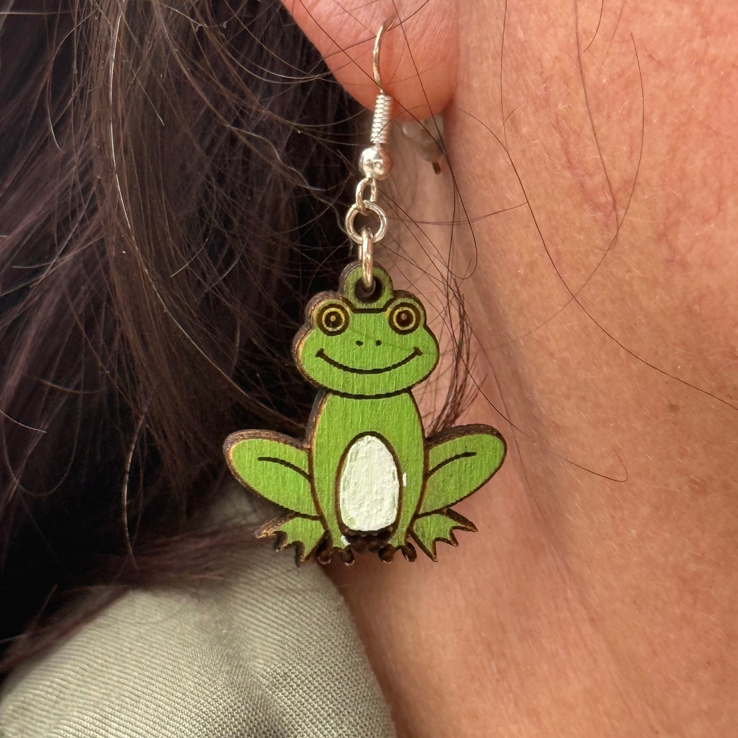Close-up of a customer wearing hand-painted green frog wooden dangle earrings. This lifestyle photo highlights the detailed artwork, lightweight design, and comfortable fit of these whimsical handmade frog earrings with hypoallergenic .925 sterling silver hooks. Perfect for frog lovers, nature enthusiasts, gardeners, amphibian fans, and anyone who enjoys playful animal jewelry.