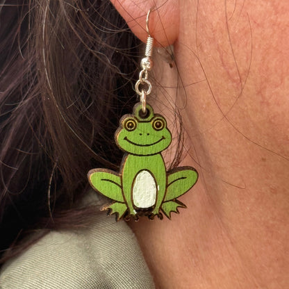 Close-up of a customer wearing hand-painted green frog wooden dangle earrings. This lifestyle photo highlights the detailed artwork, lightweight design, and comfortable fit of these whimsical handmade frog earrings with hypoallergenic .925 sterling silver hooks. Perfect for frog lovers, nature enthusiasts, gardeners, amphibian fans, and anyone who enjoys playful animal jewelry.