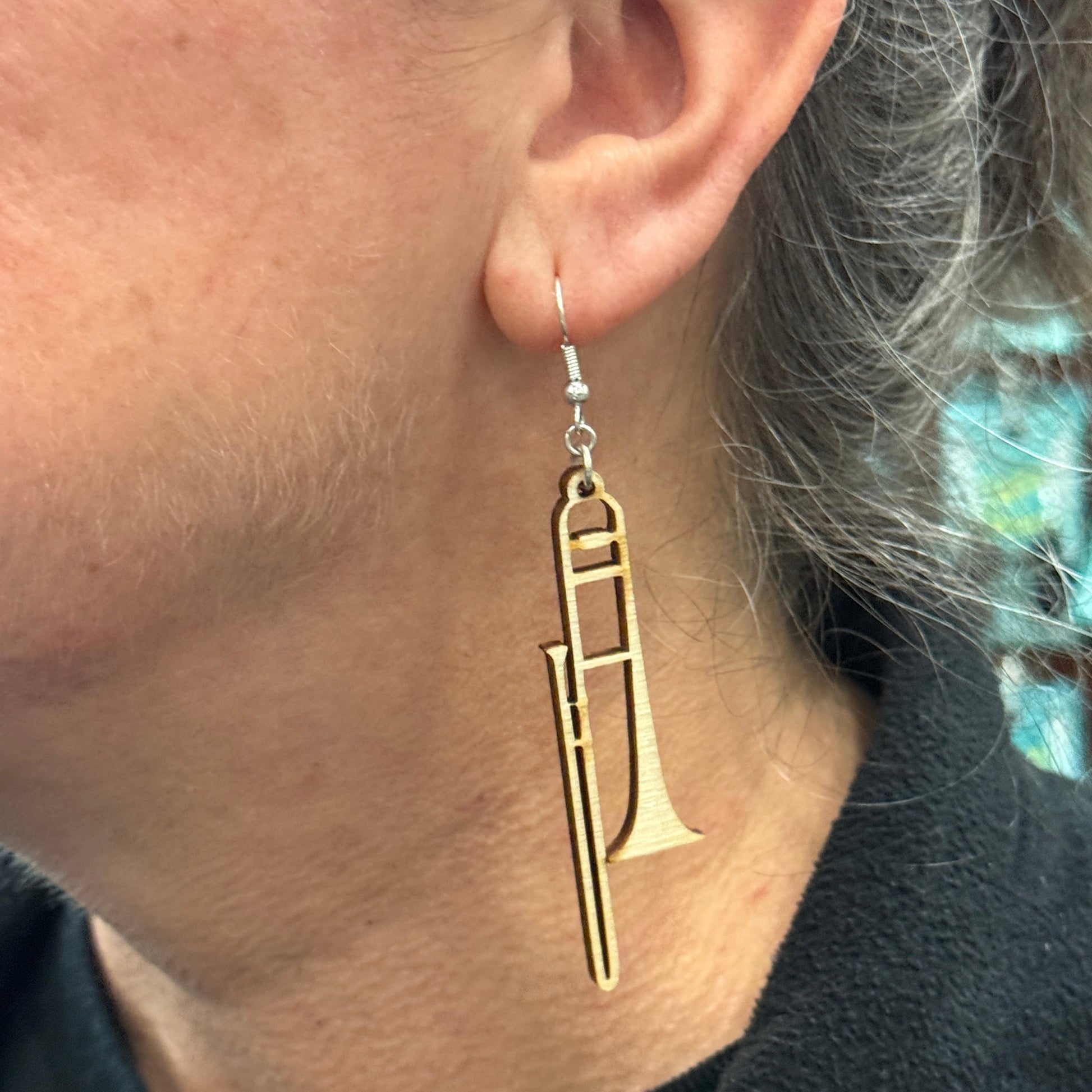 Close-up of handmade wooden trombone dangle earring worn on hypoallergenic .925 sterling silver hooks. Lightweight musician jewelry showing the size and fit, perfect for trombone players, band members, music teachers, and orchestra enthusiasts.
