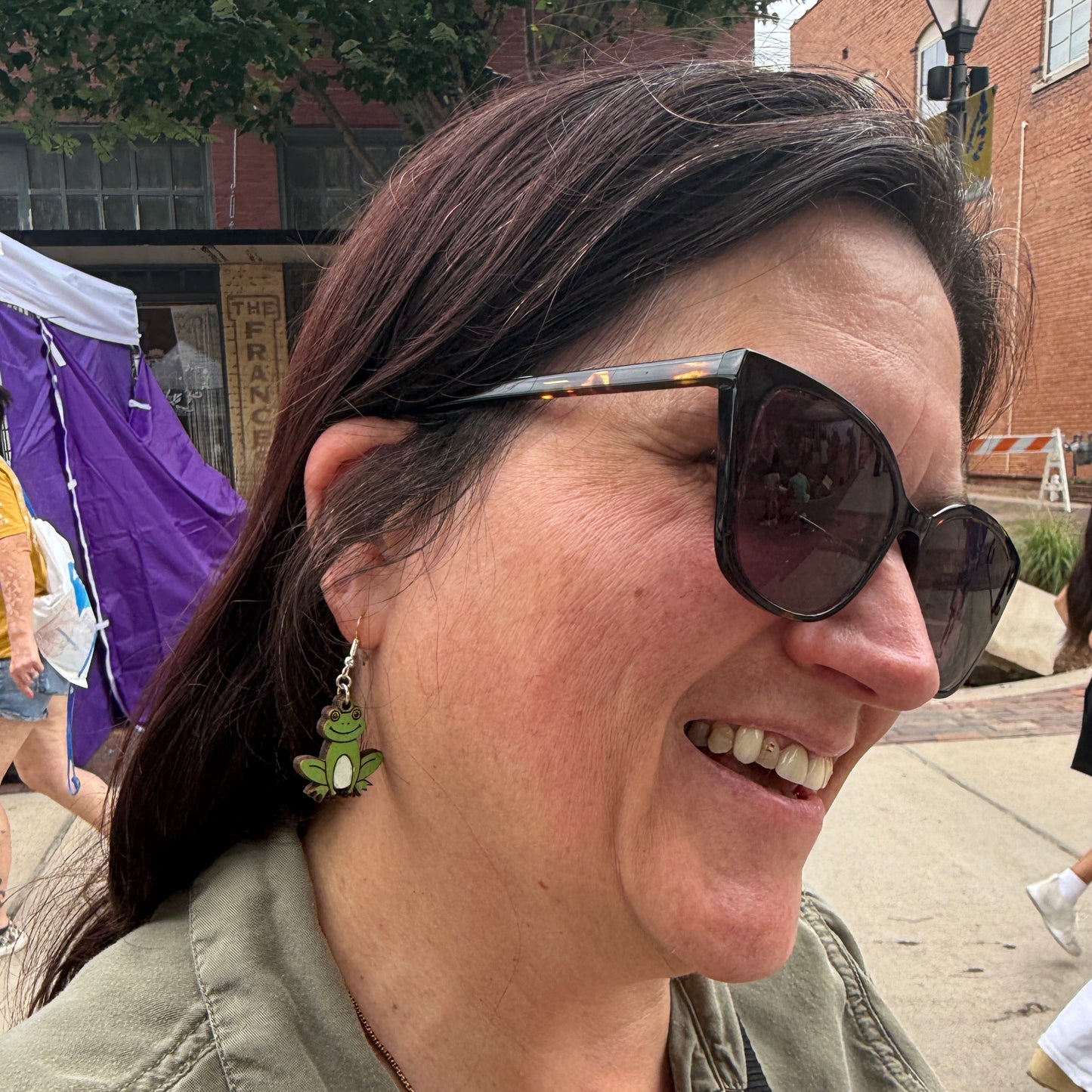Customer wearing hand-painted green frog wooden dangle earrings at an outdoor art festival. This lifestyle photo showcases the size, movement, and everyday wearability of these lightweight handmade frog earrings with hypoallergenic .925 sterling silver hooks. A fun accessory for frog lovers, wildlife enthusiasts, cottagecore fashion, and nature-inspired outfits.