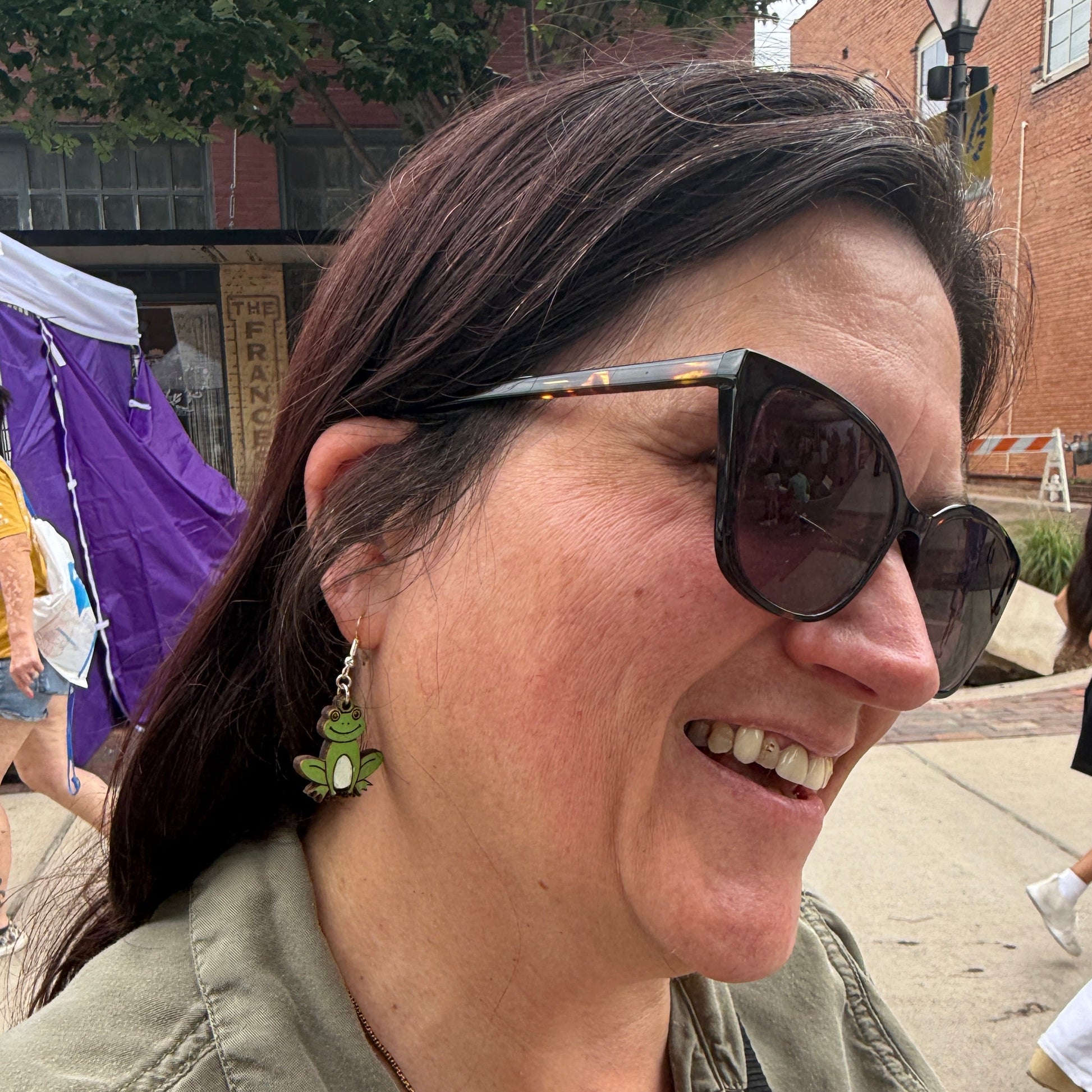 Customer wearing hand-painted green frog wooden dangle earrings at an outdoor art festival. This lifestyle photo showcases the size, movement, and everyday wearability of these lightweight handmade frog earrings with hypoallergenic .925 sterling silver hooks. A fun accessory for frog lovers, wildlife enthusiasts, cottagecore fashion, and nature-inspired outfits.