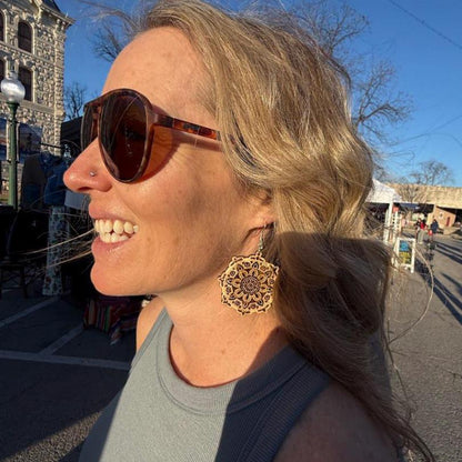 Customer wearing intricate laser-cut wooden mandala dangle earrings at an outdoor market. This lifestyle photo shows the elegant size, natural wood finish, and detailed floral mandala pattern of these lightweight earrings with hypoallergenic .925 sterling silver hooks. Perfect for boho fashion, festivals, and everyday wear.