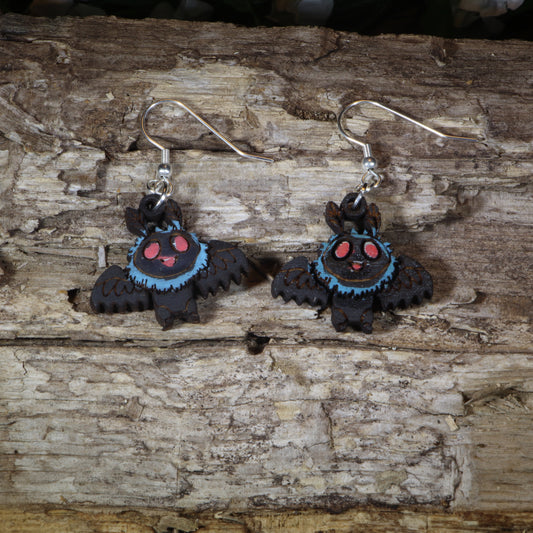 Hand-painted wooden cute Mothman dangle earrings featuring an adorable winged cryptid with glowing red eyes, hypoallergenic sterling silver hooks, and lightweight cryptid mythology jewelry for folklore fans and paranormal enthusiasts by Cate's Concepts.