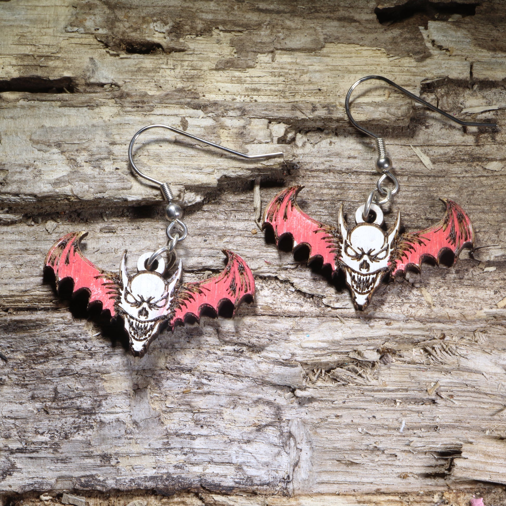 Demon skull bat wooden dangle earrings featuring a sinister horned skull with outstretched bat wings and gothic horror details. Lightweight hand-painted wooden earrings with hypoallergenic .925 sterling silver hooks, perfect for horror fans, gothic fashion, Halloween, occult-inspired style, and spooky statement jewelry.
