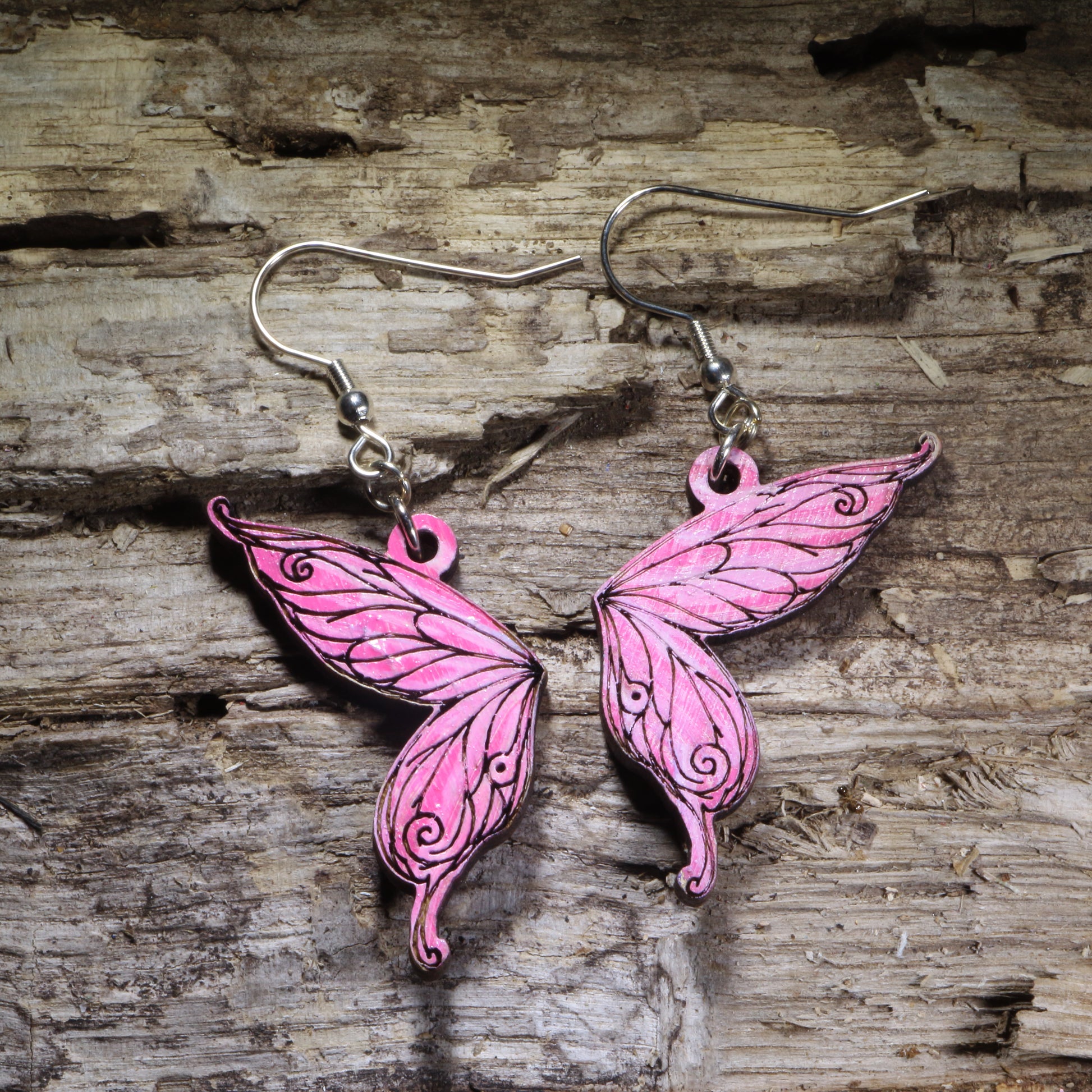 Fairy wing wooden dangle earrings featuring detailed hand-painted wings in pink with whimsical swirling accents. Lightweight fantasy earrings with hypoallergenic .925 sterling silver hooks, perfect for fairy lovers, fantasy fans, cottagecore style, renaissance festival outfits, and magical jewelry gifts.