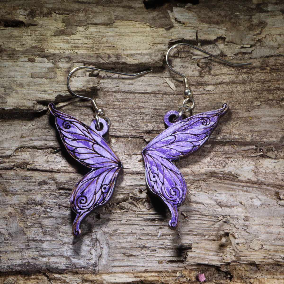 Fairy wing wooden dangle earrings featuring detailed hand-painted wings in purple with whimsical swirling accents. Lightweight fantasy earrings with hypoallergenic .925 sterling silver hooks, perfect for fairy lovers, fantasy fans, cottagecore style, renaissance festival outfits, and magical jewelry gifts.