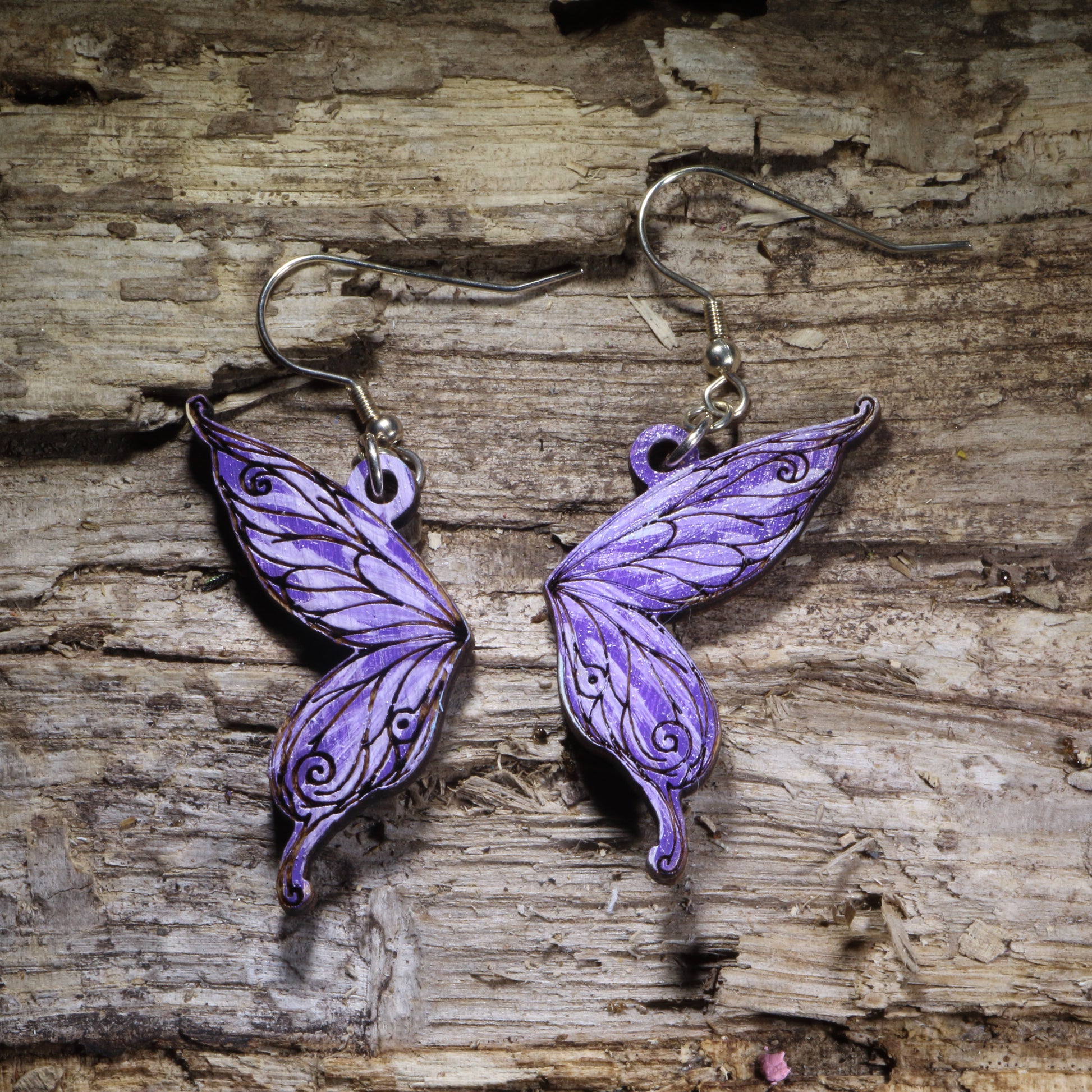 Fairy wing wooden dangle earrings featuring detailed hand-painted wings in purple with whimsical swirling accents. Lightweight fantasy earrings with hypoallergenic .925 sterling silver hooks, perfect for fairy lovers, fantasy fans, cottagecore style, renaissance festival outfits, and magical jewelry gifts.