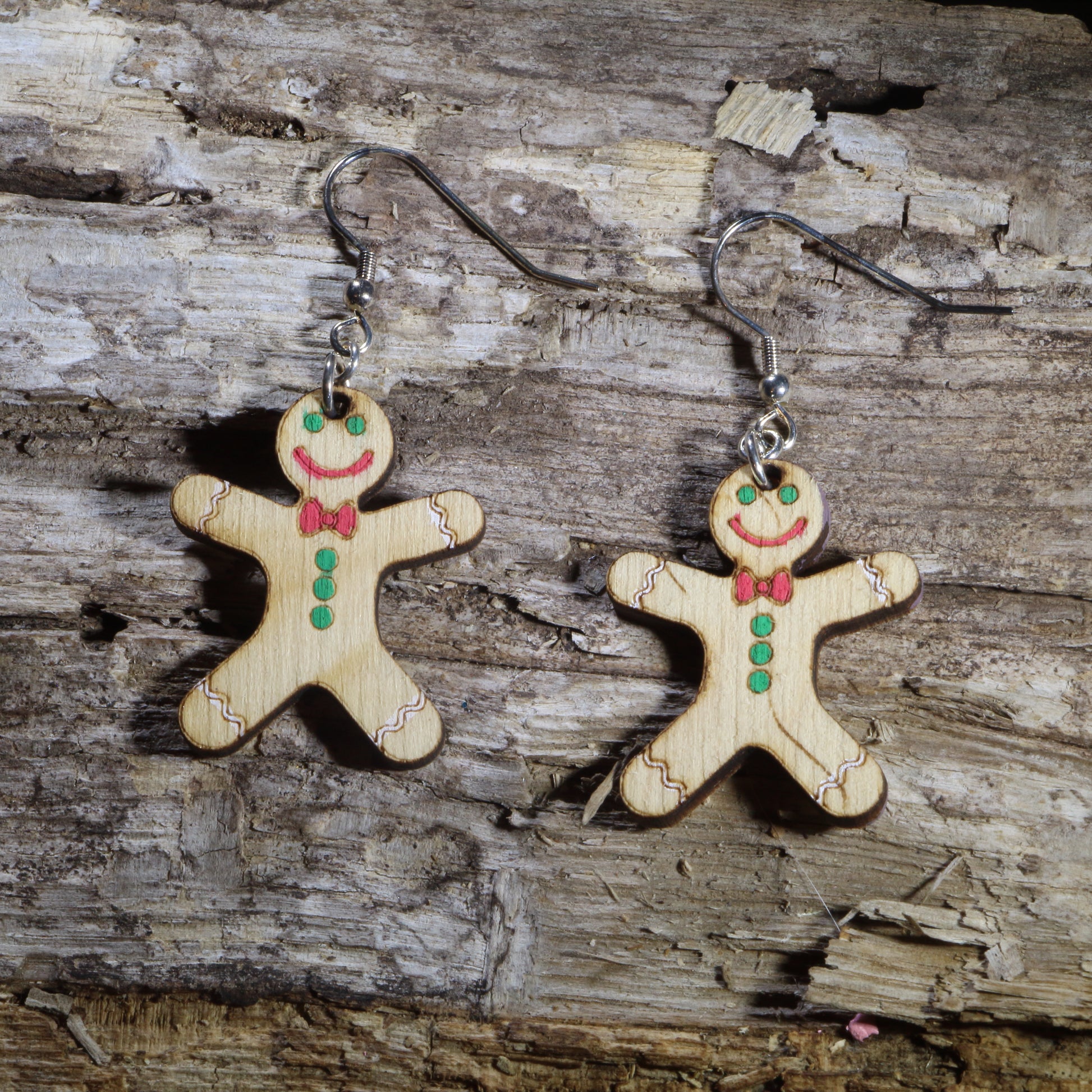 Hand-painted wooden gingerbread man dangle earrings featuring smiling gingerbread cookies decorated with green candy buttons, a red bow tie, pink icing details, and cheerful holiday accents. Crafted from lightweight wood with hypoallergenic .925 sterling silver hooks, these festive Christmas earrings are perfect for holiday baking, Christmas parties, cookie exchanges, winter celebrations, stocking stuffers, and anyone who loves classic gingerbread holiday jewelry.