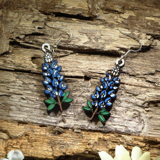 Hand-painted wooden Texas Bluebonnet dangle earrings featuring the iconic blue state flower of Texas with vibrant floral details. Crafted from lightweight wood with hypoallergenic .925 sterling silver hooks, these artisan earrings are perfect for Texas pride, wildflower lovers, spring festivals, Bluebonnet season, nature enthusiasts, travel souvenirs, and anyone who loves colorful botanical jewelry inspired by the Lone Star State.