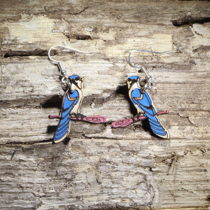 Hand-painted wooden blue jay dangle earrings featuring two blue jays perched on pink tree branches with hypoallergenic .925 sterling silver hooks. Lightweight bird lover jewelry inspired by backyard songbirds, perfect for nature enthusiasts, birdwatchers, and wildlife gifts.