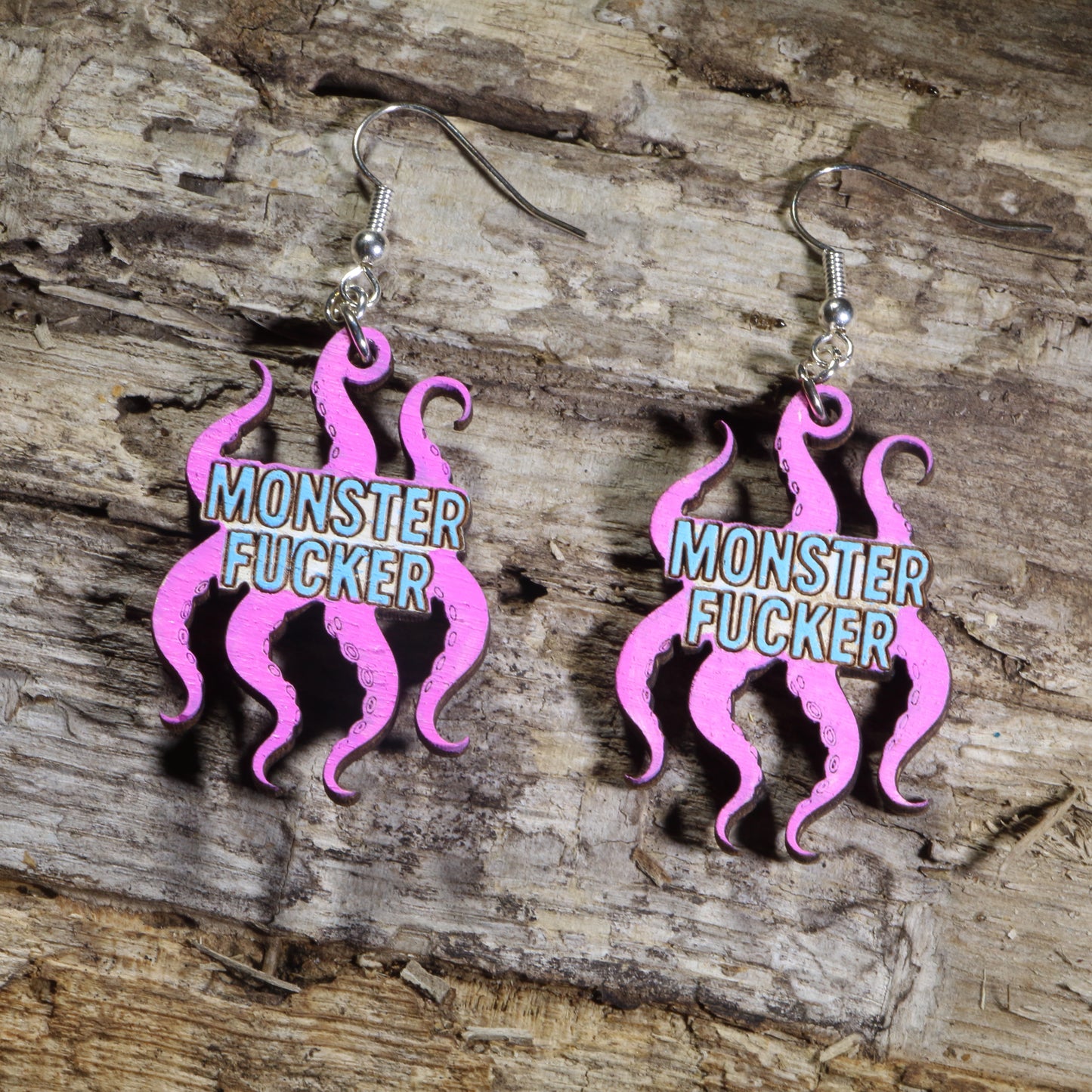 Hand-painted wooden octopus dangle earrings featuring bright pink tentacles and the phrase "Monster Fucker" in blue lettering on hypoallergenic .925 sterling silver hooks. Lightweight fantasy-inspired statement jewelry with a humorous adult design for monster romance fans and book lovers.