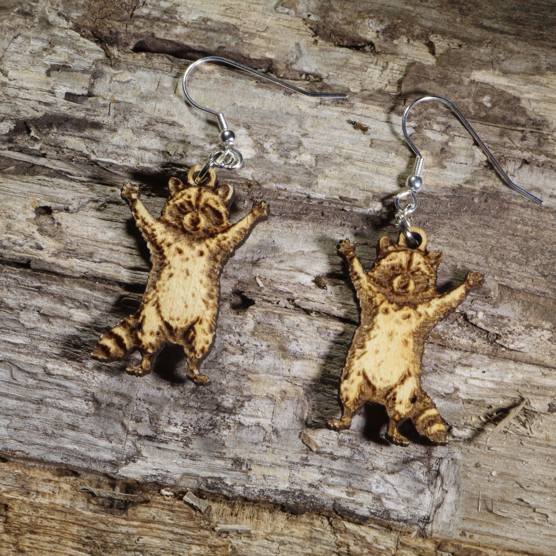 Hand-painted wooden raccoon dangle earrings featuring an adorable raccoon with raised paws and engraved fur details on hypoallergenic .925 sterling silver hooks. Lightweight woodland animal jewelry perfect for raccoon lovers, wildlife enthusiasts, and nature-inspired accessories
