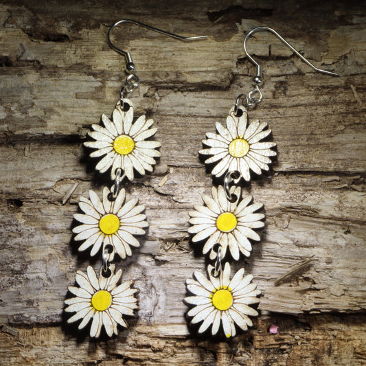 Handcrafted daisy chain wooden dangle earrings featuring three hand-painted white daisies with bright yellow centers linked together for graceful movement. Lightweight laser-cut floral earrings with hypoallergenic .925 sterling silver hooks, perfect for cottagecore fashion, wildflower lovers, spring and summer outfits, botanical jewelry, garden enthusiasts, and nature-inspired accessories
