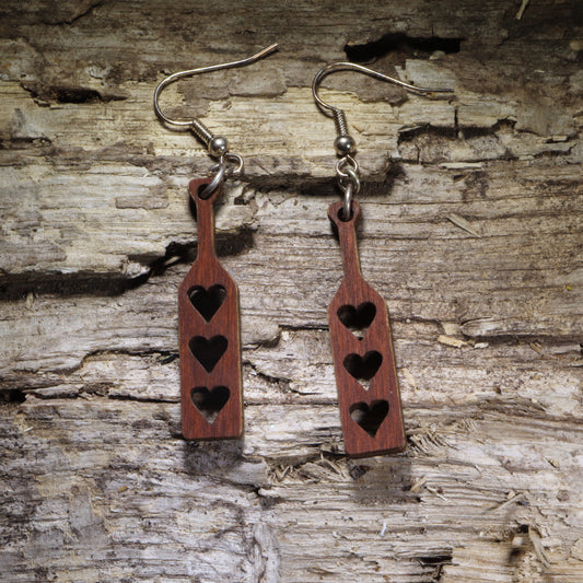 Handcrafted wooden spanking paddle dangle earrings featuring three heart-shaped cutouts in a classic adult paddle design. Lightweight laser-cut wooden earrings with hypoallergenic .925 sterling silver hooks, perfect for BDSM enthusiasts, kink fashion, fetish events, adult humor, playful gifts, and handmade novelty jewelry.