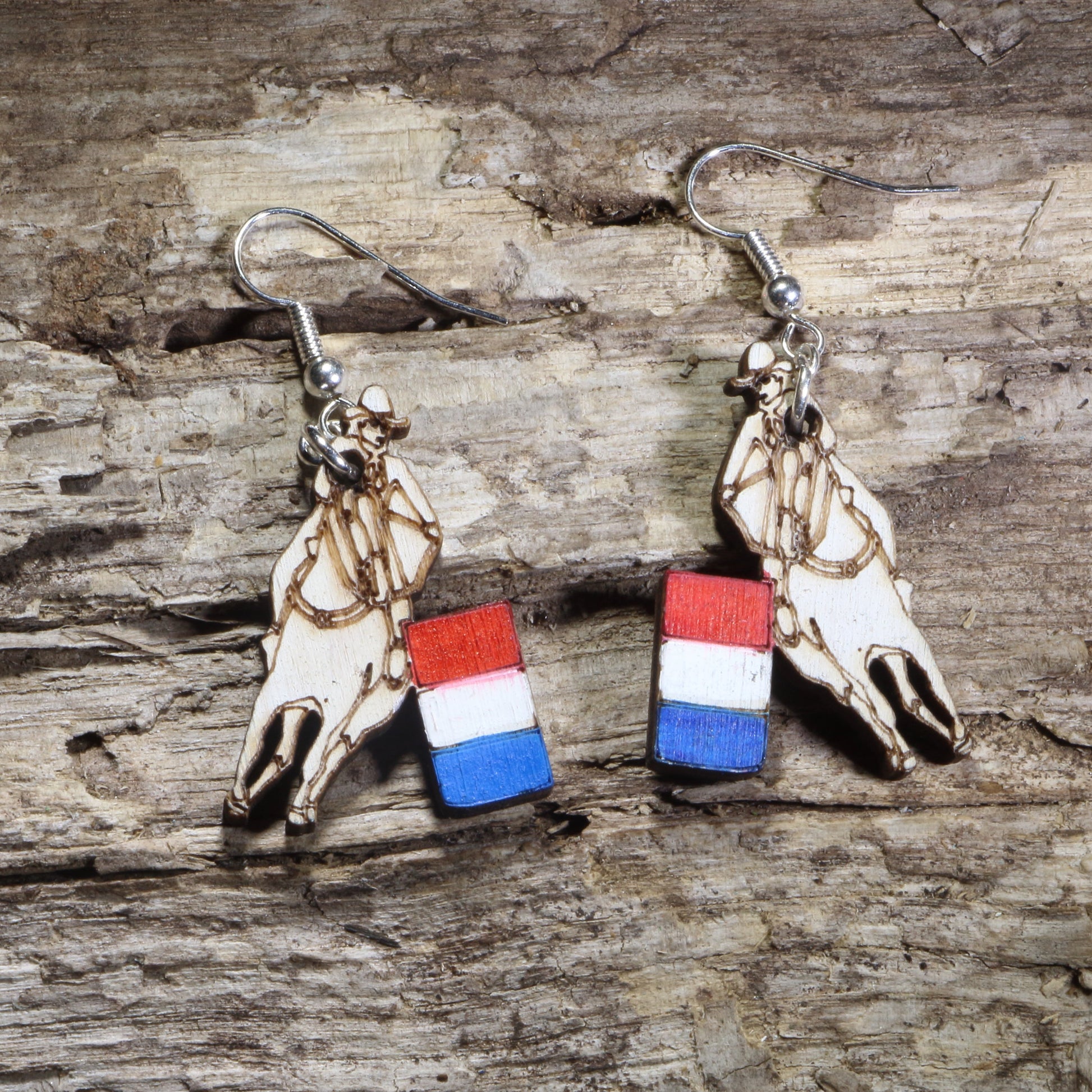 Handmade barrel racing wooden dangle earrings featuring a laser-cut western saddle with a red, white, and blue racing barrel charm. Lightweight patriotic rodeo earrings crafted from wood with hypoallergenic .925 sterling silver hooks, perfect for barrel racers, horse lovers, equestrians, western fashion, and country lifestyle enthusiasts.
