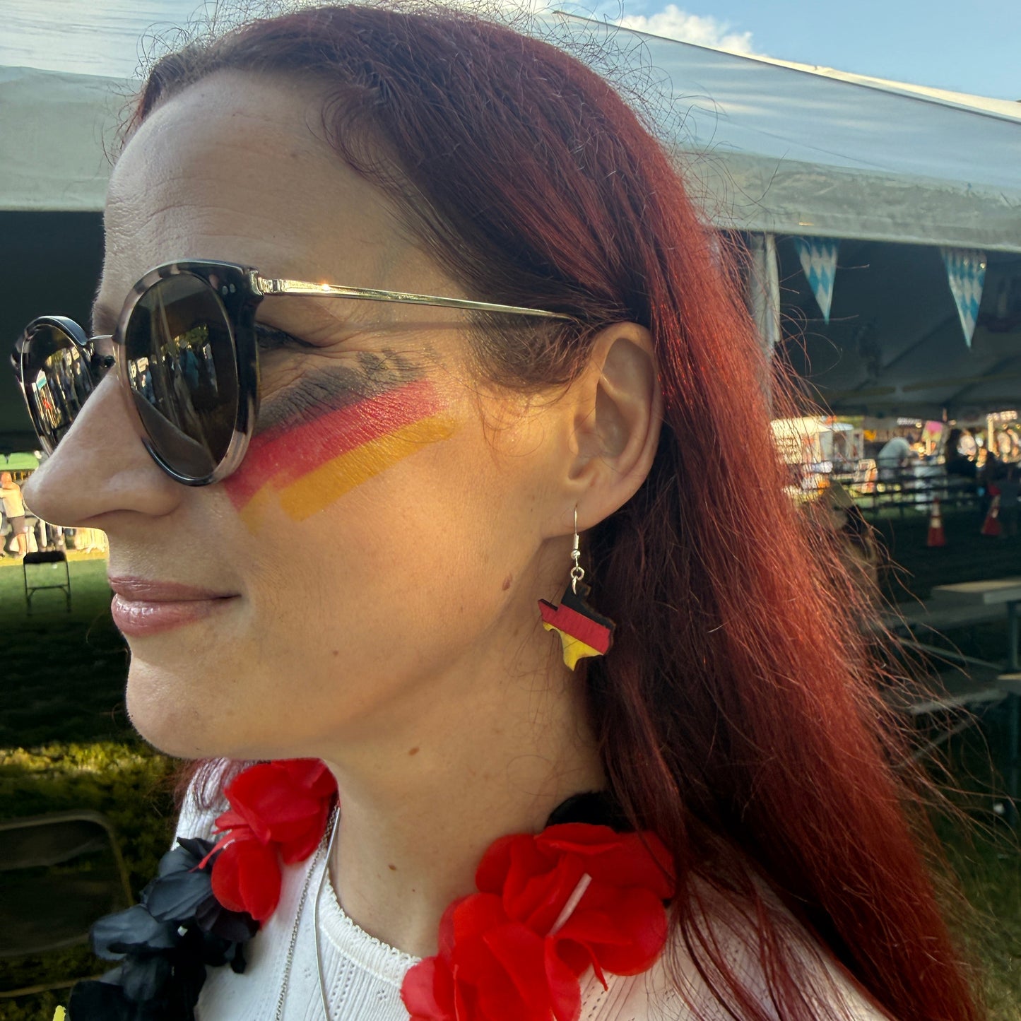 Woman wearing hand-painted wooden Germany state-shaped dangle earrings with German flag colors at an outdoor cultural festival, lightweight heritage jewelry by Cate’s Concepts