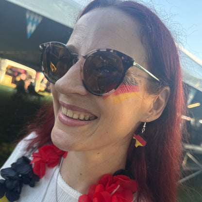 Smiling woman modeling hand-painted wooden Texas-shaped dangle earrings with German flag colors at a festival celebration, lightweight Texas heritage jewelry by Cate’s Concepts