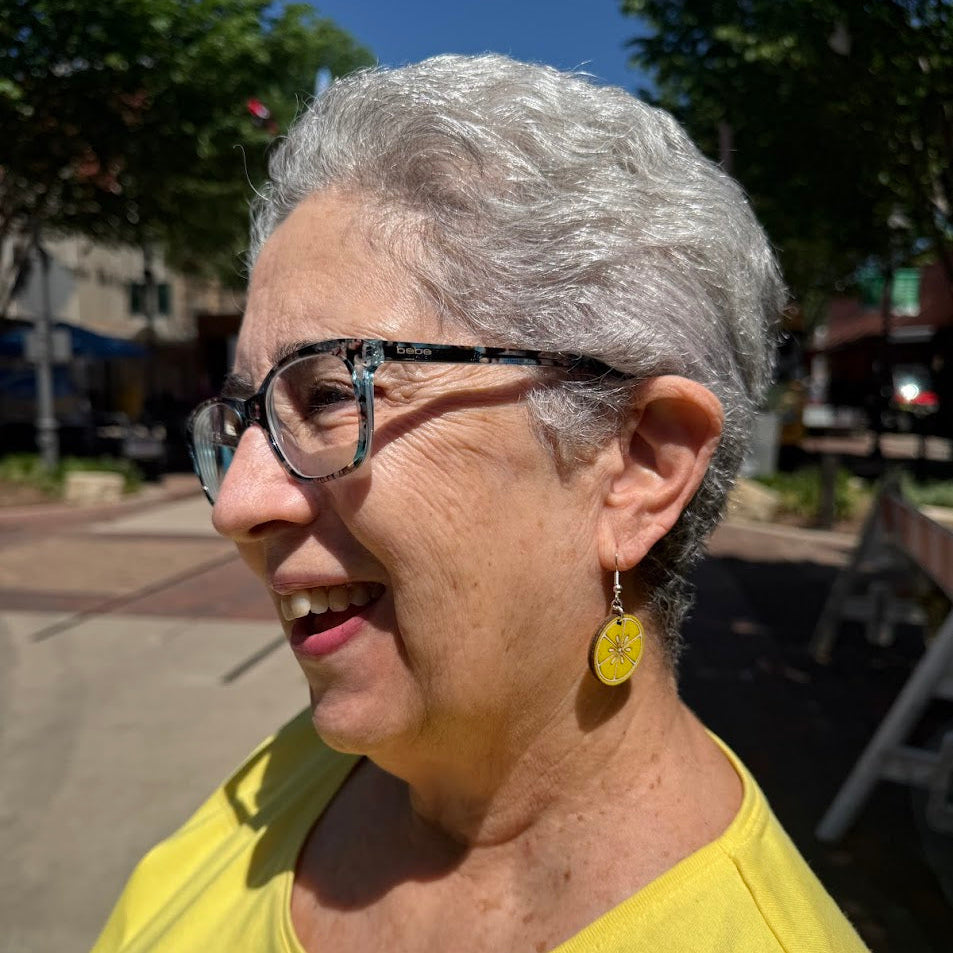 Smiling woman wearing hand-painted wooden lemon slice dangle earrings at an outdoor festival, lightweight citrus fruit summer jewelry by Cate’s Concepts