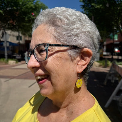 Smiling woman wearing hand-painted wooden lemon slice dangle earrings at an outdoor festival, lightweight citrus fruit summer jewelry by Cate’s Concepts