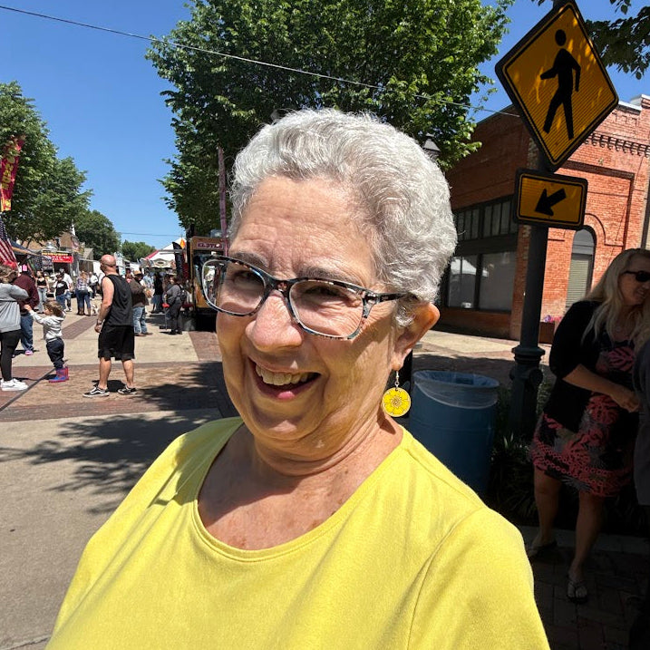 Woman modeling hand-painted wooden lemon slice dangle earrings at a sunny street festival, lightweight summer fruit jewelry by Cate’s Concepts