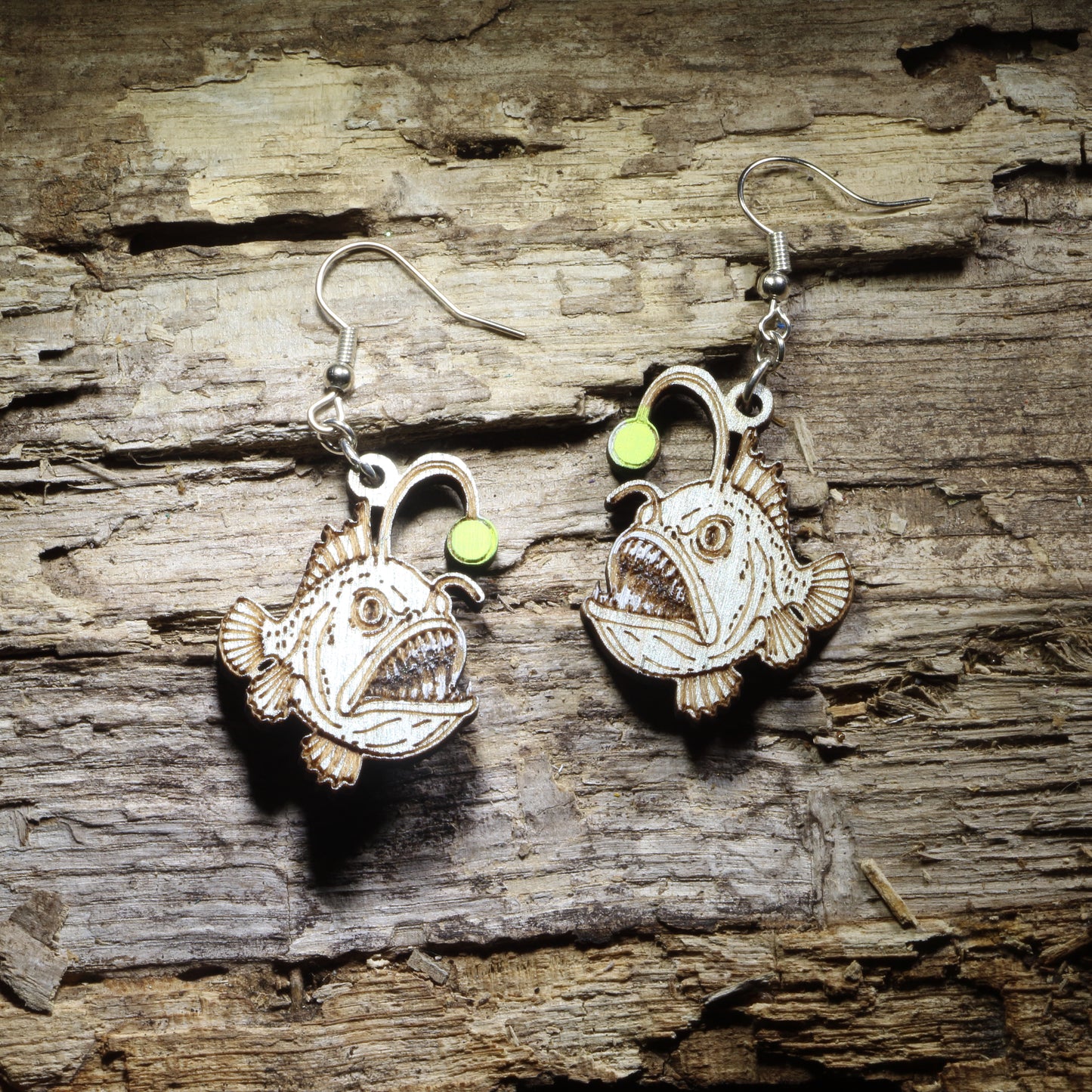 Pair of hand-painted anglerfish wooden dangle earrings featuring detailed deep-sea fish with bright green bioluminescent lures on rustic wood