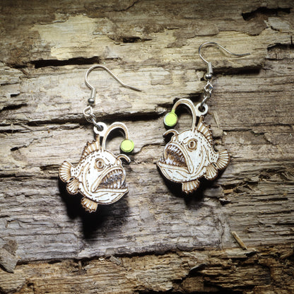 Pair of hand-painted anglerfish wooden dangle earrings featuring detailed deep-sea fish with bright green bioluminescent lures on rustic wood