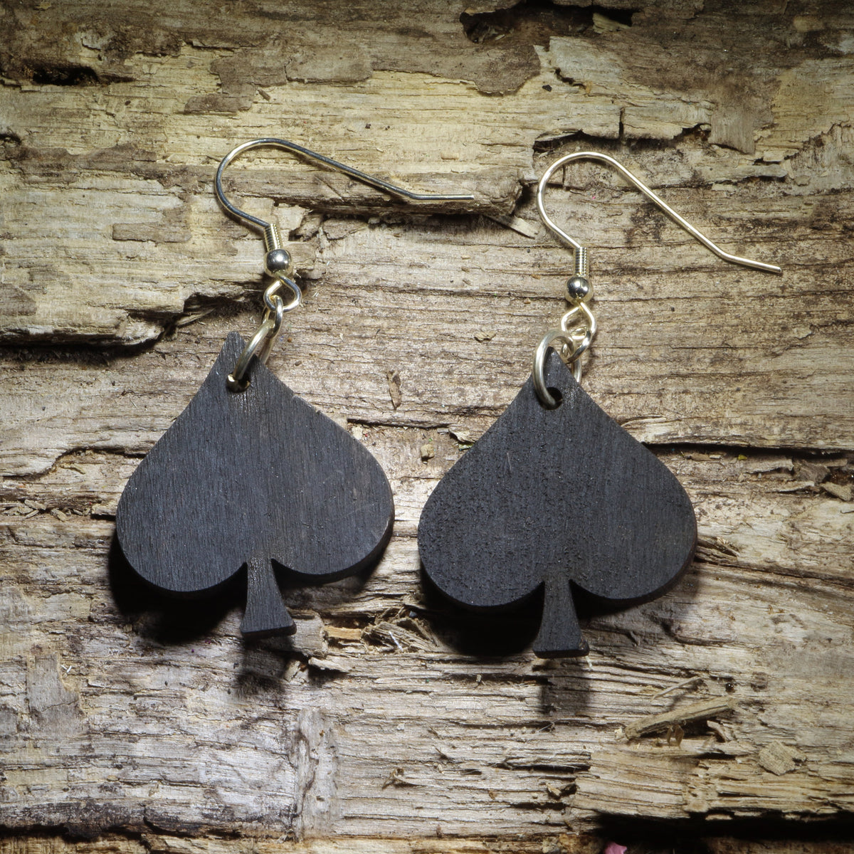 Pair of hand-painted black spade wooden dangle earrings featuring a bold playing card suit design displayed on rustic weathered wood