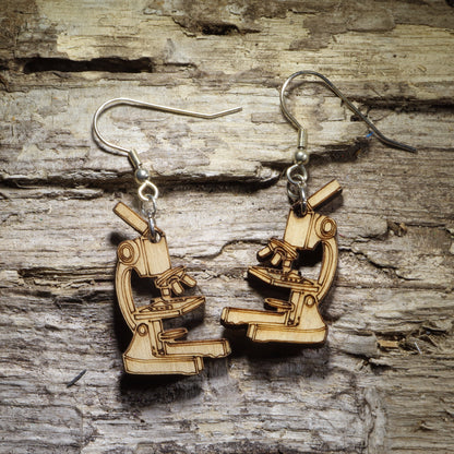 Pair of natural wood microscope dangle earrings featuring detailed scientific laboratory microscope designs, displayed on rustic weathered wood