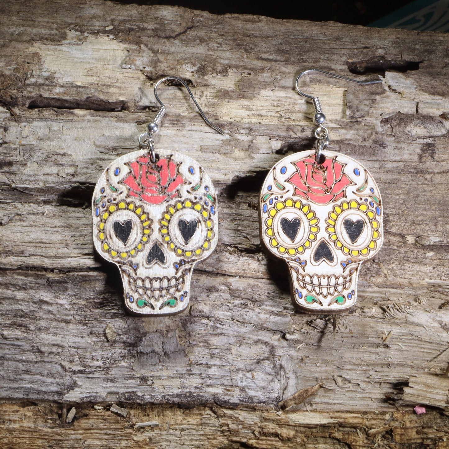 Hand-painted wooden sugar skull dangle earrings inspired by Día de los Muertos, featuring white skulls with heart-shaped eyes, yellow floral accents, and red roses on hypoallergenic .925 sterling silver hooks. Lightweight gothic and Day of the Dead jewelry perfect for Halloween, Mexican folk art lovers, and unique statement style.