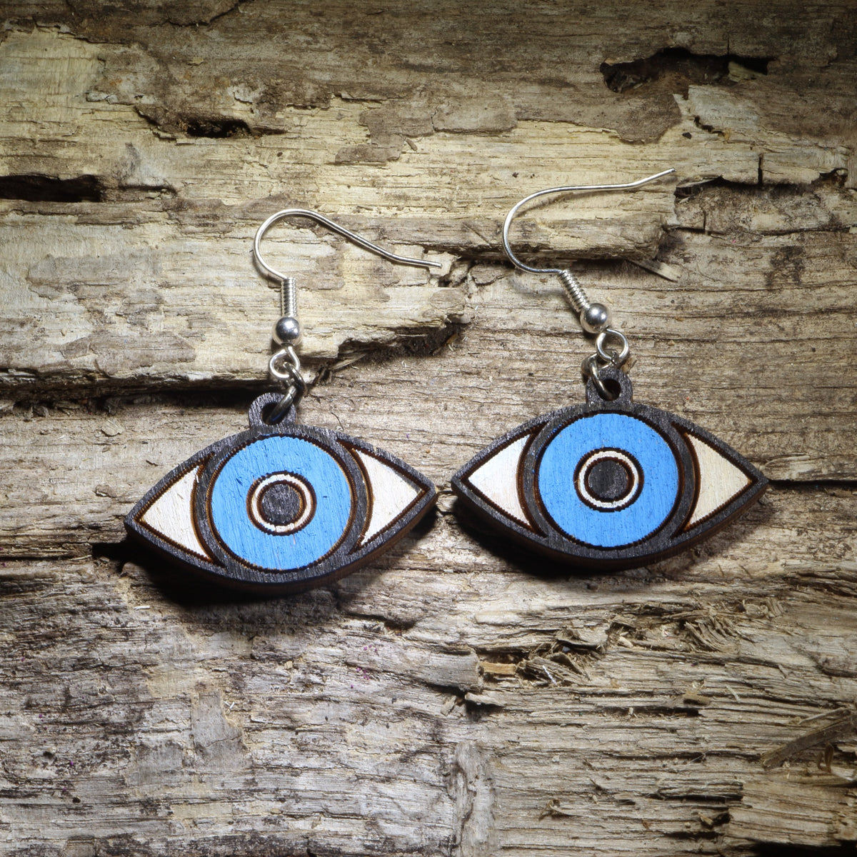 Blue and white evil eye wooden dangle earrings with bright blue circular centers, black and white eye details, and silver hooks on a weathered wood background