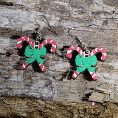 Hand-painted wooden crossed candy cane dangle earrings featuring two festive red and white candy canes tied together with a bright green holiday bow. Crafted from lightweight wood with hypoallergenic .925 sterling silver hooks, these cheerful Christmas earrings are perfect for holiday parties, Christmas celebrations, winter festivals, stocking stuffers, cookie exchanges, and anyone who loves classic Christmas candy and festive holiday jewelry.