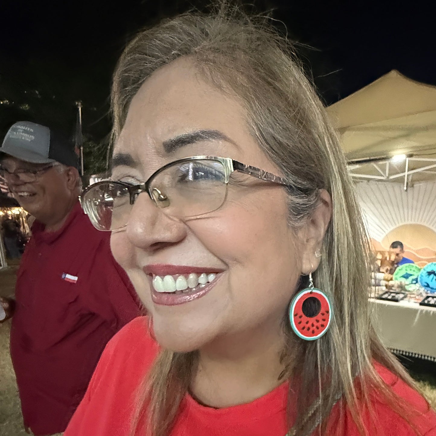 Woman wearing hand-painted wooden watermelon slice dangle earrings at an outdoor market, showcasing the colorful red, green, and white watermelon design. This lifestyle photo demonstrates the size and lightweight comfort of these handmade wooden earrings with hypoallergenic .925 sterling silver hooks, perfect for summer festivals, vacations, picnics, and casual everyday style.