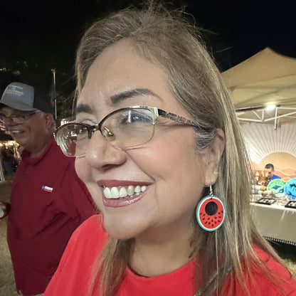 Woman wearing hand-painted wooden watermelon slice dangle earrings at an outdoor market, showcasing the colorful red, green, and white watermelon design. This lifestyle photo demonstrates the size and lightweight comfort of these handmade wooden earrings with hypoallergenic .925 sterling silver hooks, perfect for summer festivals, vacations, picnics, and casual everyday style.